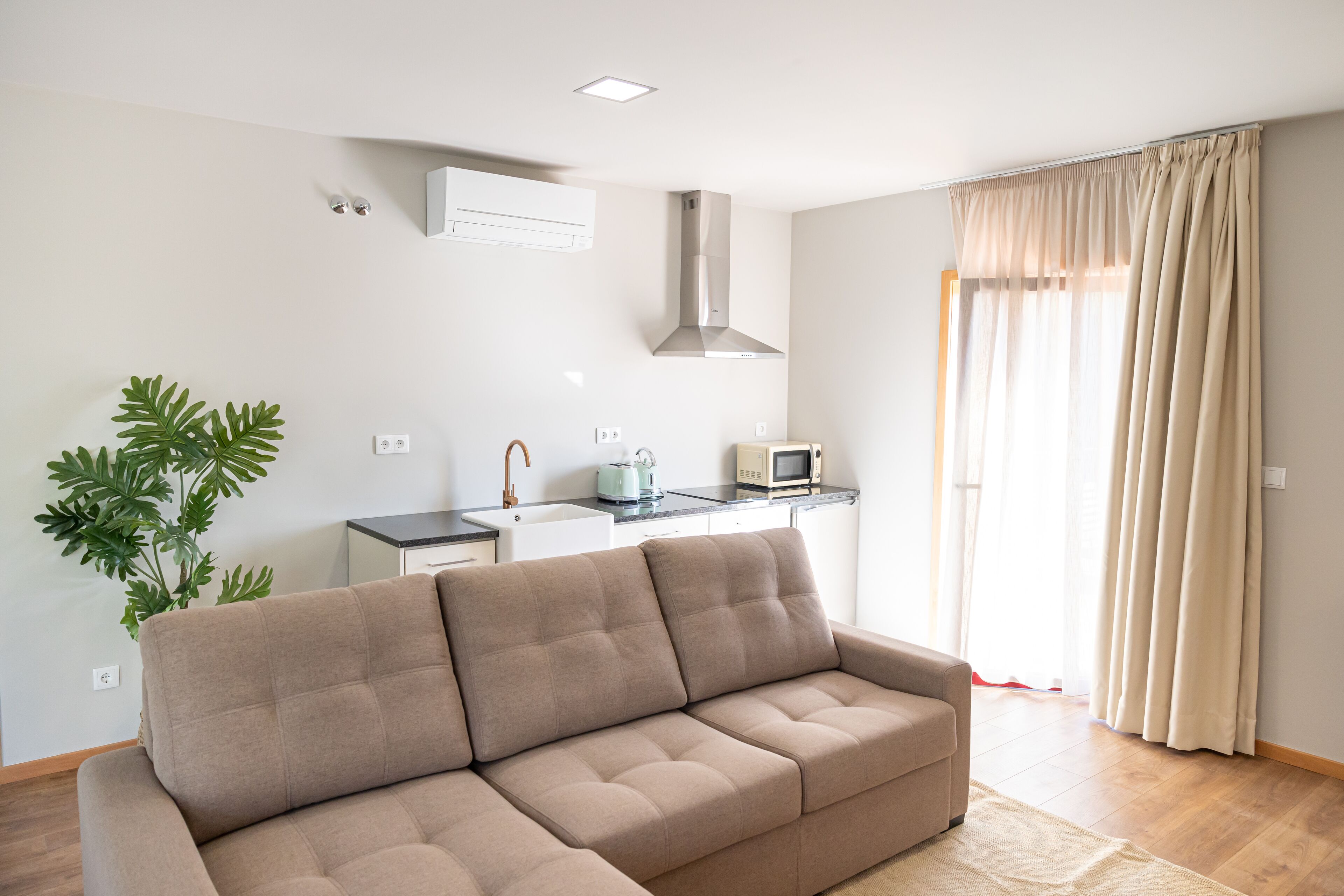 A modern living room with a beige sofa, a kitchen area with a microwave, and a large window with curtains.