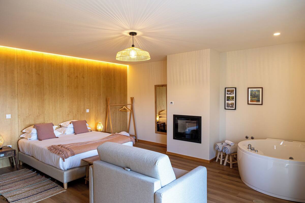 A modern bedroom with a large bed, a sitting area, a bathtub, and a fireplace.
