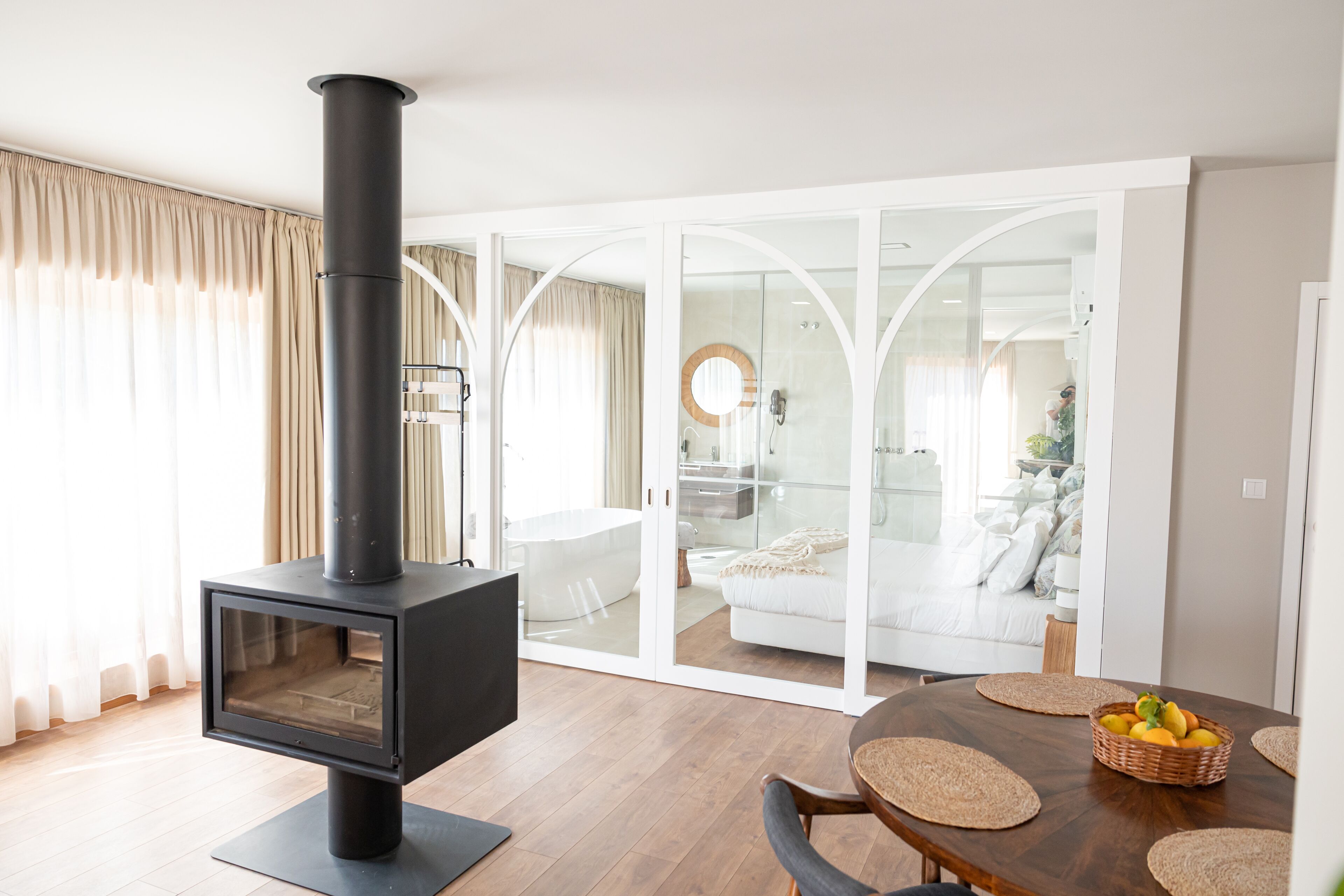 A modern living room with a wood-burning stove, a dining area with a round table and chairs, and a glass-enclosed bathroom with a bathtub.