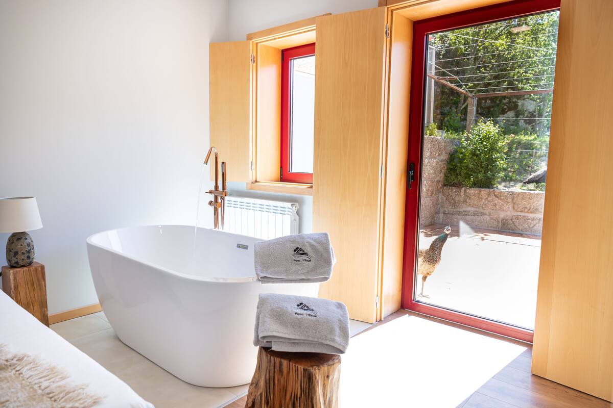 A modern bathroom with a freestanding bathtub, a wooden stool with towels, and a view of a peacock outside.