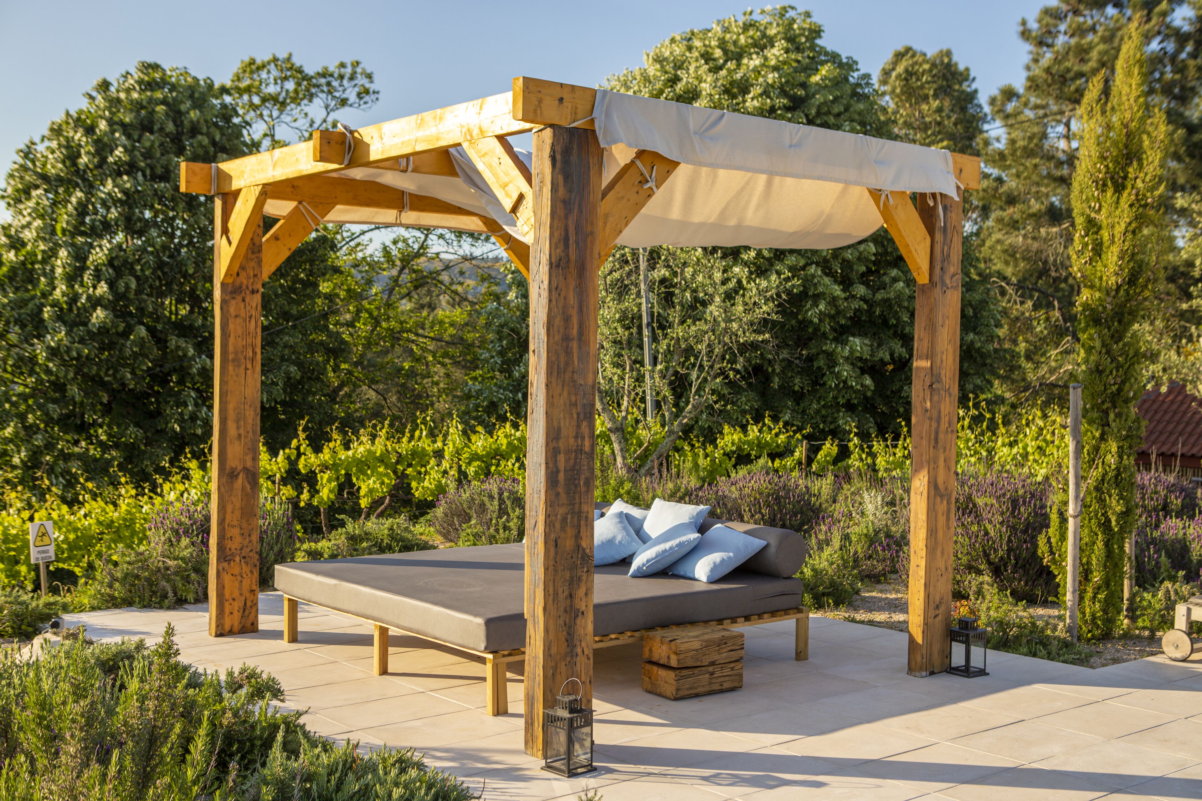 Een houten pergola met een bed eronder, omgeven door een tuin met diverse planten en bomen.