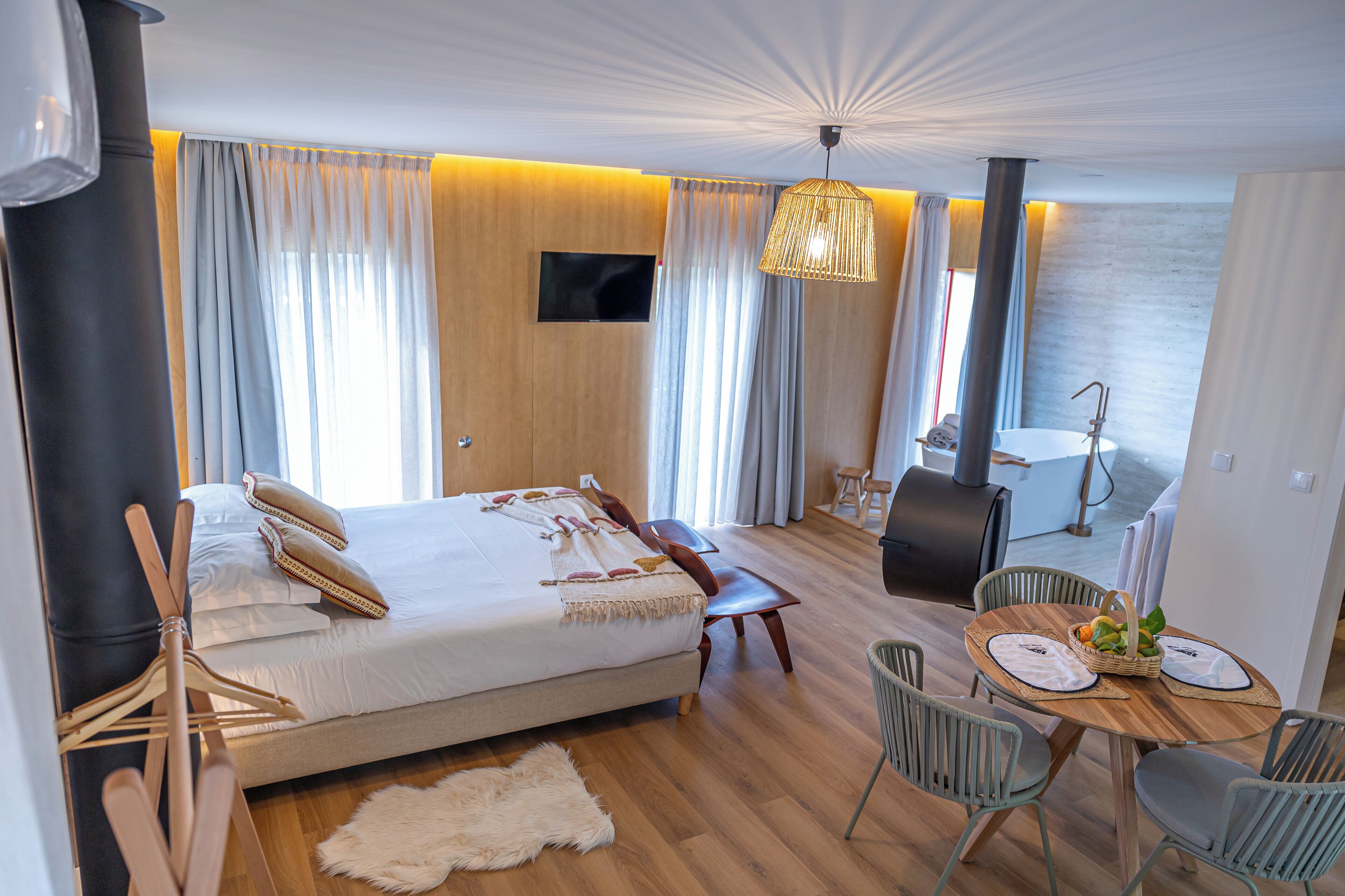 A modern hotel room with a bed, a dining table with chairs, a TV, and a view of the sea.