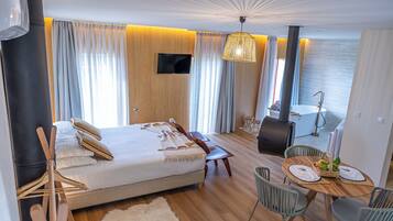 A modern hotel room with a bed, a dining table with chairs, a TV, and a view of the sea.