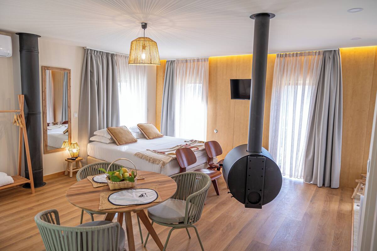 A modern hotel room with a bed, a dining table with chairs, a fireplace, and large windows with curtains.