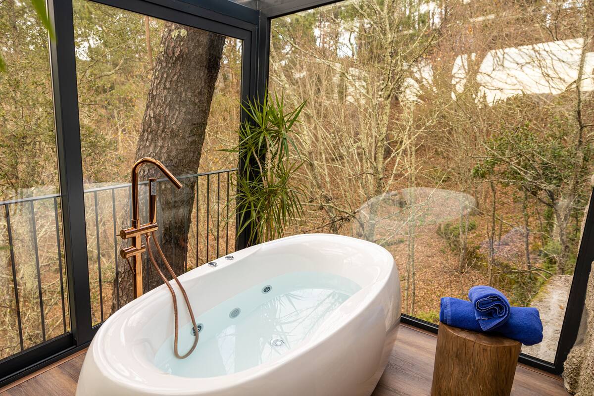A modern bathroom with a freestanding bathtub, a wooden stool, and a view of a forest through large windows.