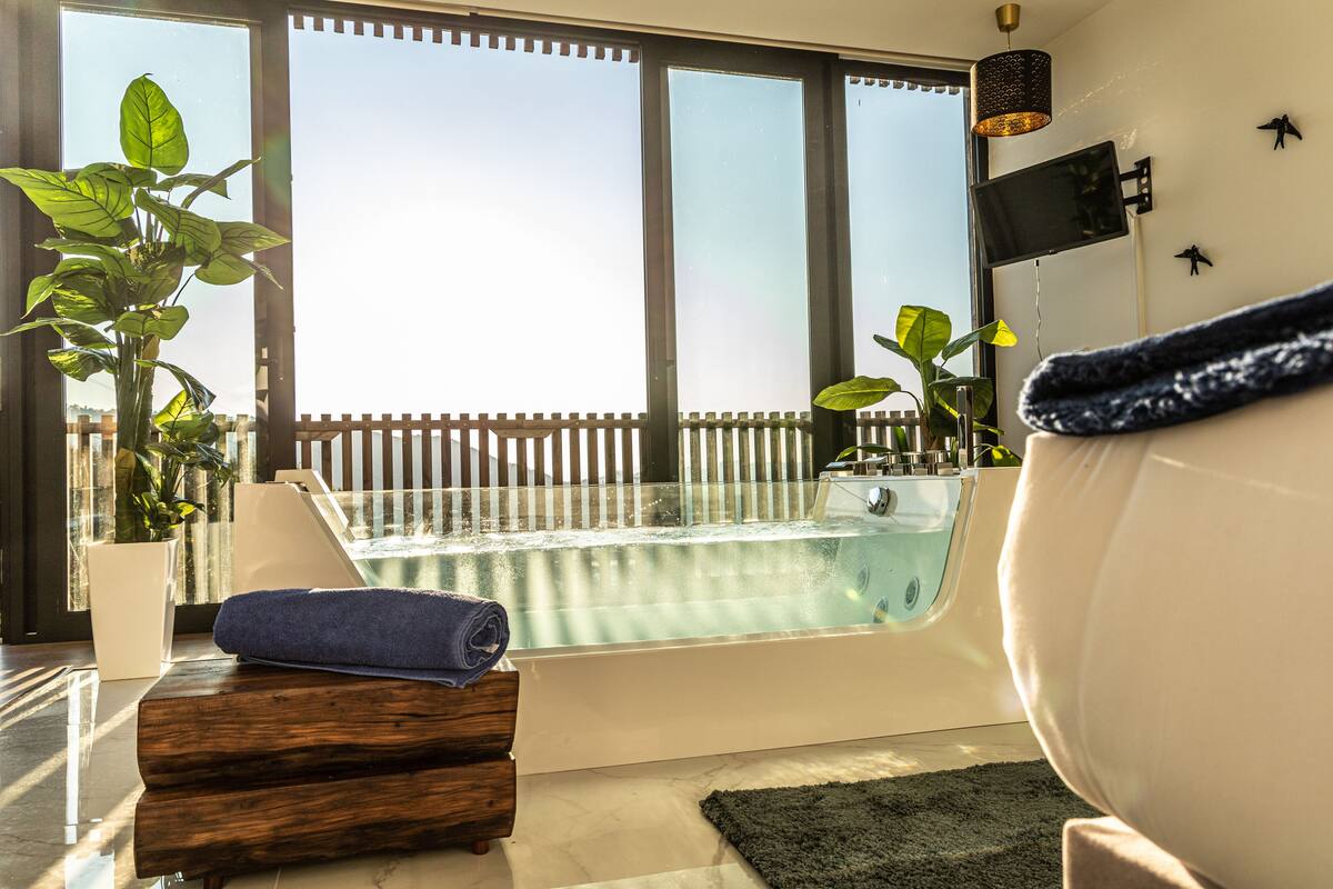 A modern bathroom with a bathtub, a potted plant, and a towel on a wooden stool.