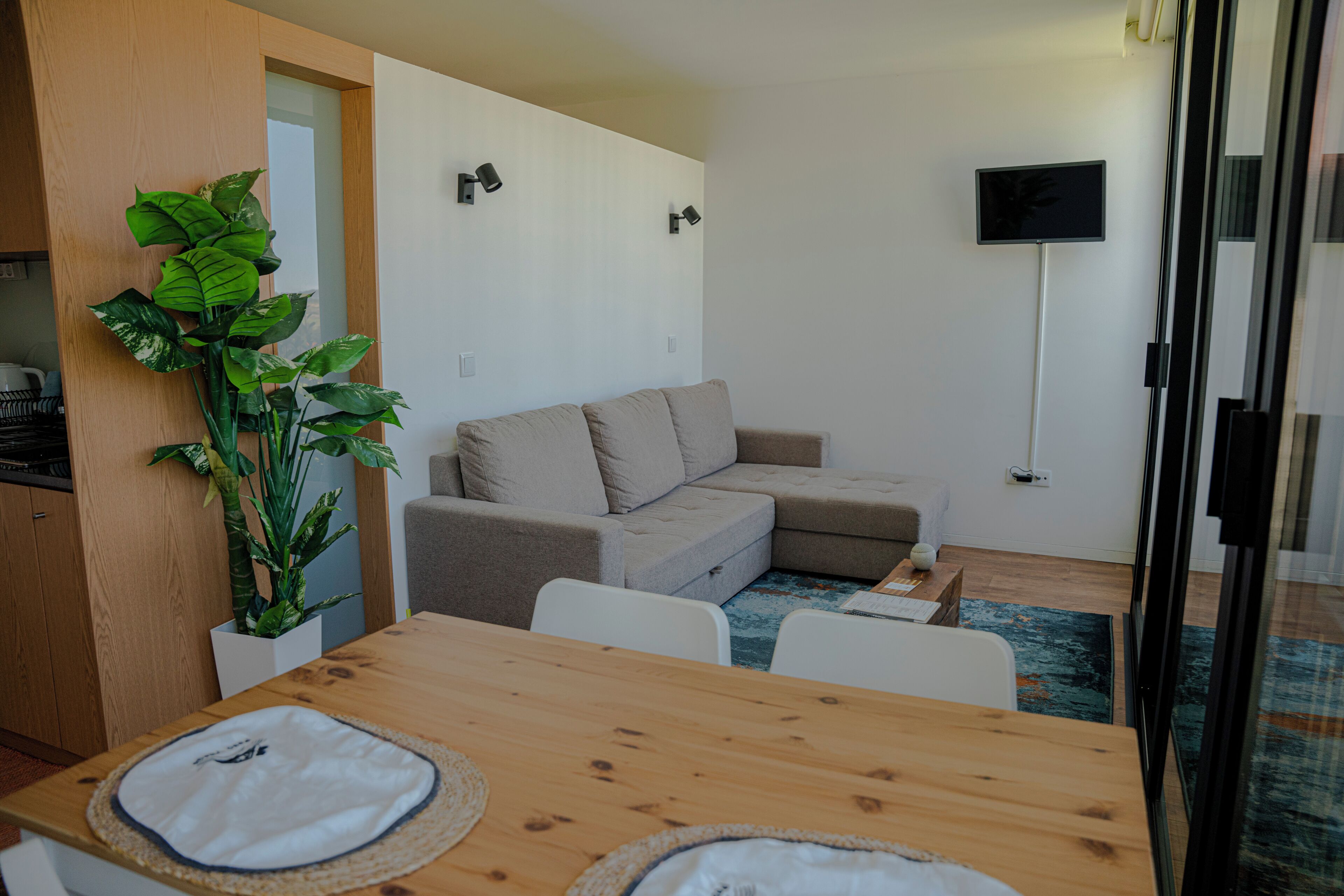 Ett modernt vardagsrum med ett matbord i trä, en beige soffa, en stor växt och en platt-TV som är monterad på väggen.