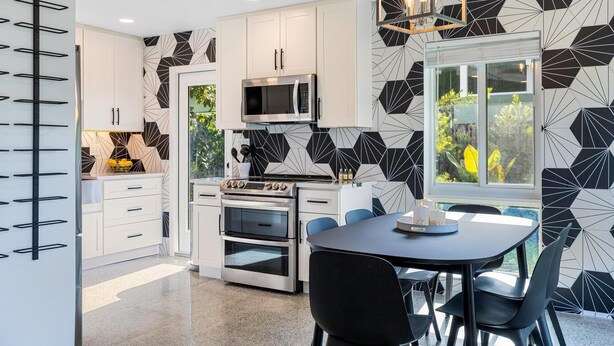 A modern kitchen with white cabinets, a built-in oven, microwave, and gas stove. There is a dining area with a table and chairs, and a window with a view of greenery outside.