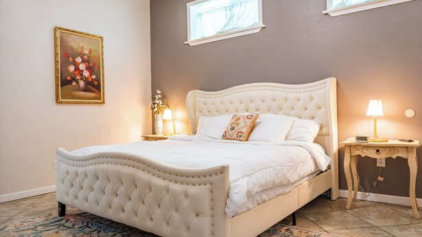 A bedroom with a tufted headboard, a floral painting, bedside lamps, and a window with curtains.