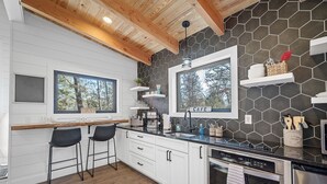 A modern kitchen with white cabinets, a black countertop, and a hexagonal tile backsplash. There are two bar stools, a large window, and a built-in oven. - Romantic Cabin - Lakeside (Jonesburg)