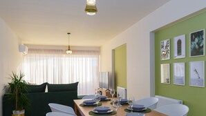Ein Essbereich mit einem Holztisch, gedeckt für vier Personen, umgeben von weißen Stühlen. In der Ecke befindet sich eine grüne Couch und ein Blumentopf.
