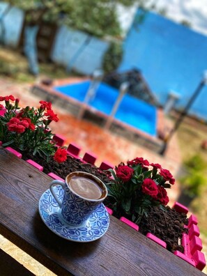 A cup of coffee on a wooden table with red flowers and a blue pool in the background. - Zeytindagi Bungalow (Edremit)