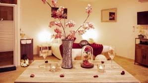 A room with a wooden table set for a romantic dinner, featuring a vase with pink flowers, a glass candle holder, and scattered rose petals.