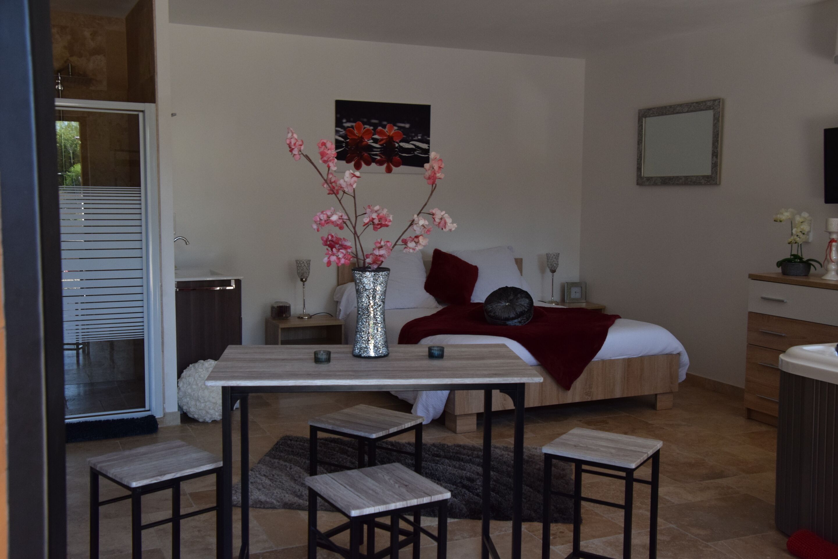 A bedroom with a bed, a bedside table, a small table with chairs, a vase with flowers, and a painting on the wall.