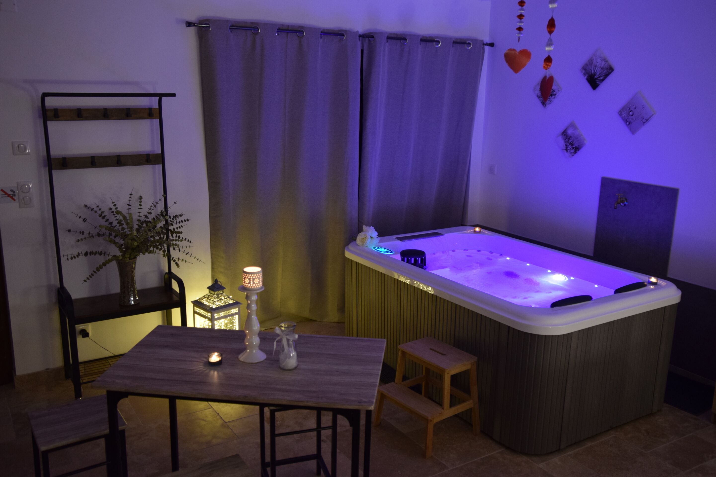 A room with a hot tub, a table with candles, a chair, a small stool, a plant, and a curtain.