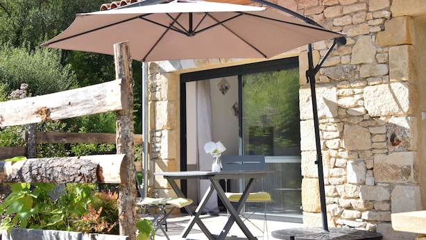 An outdoor patio with a table and chairs, a large umbrella, and a stone building with a wooden fence in the background.