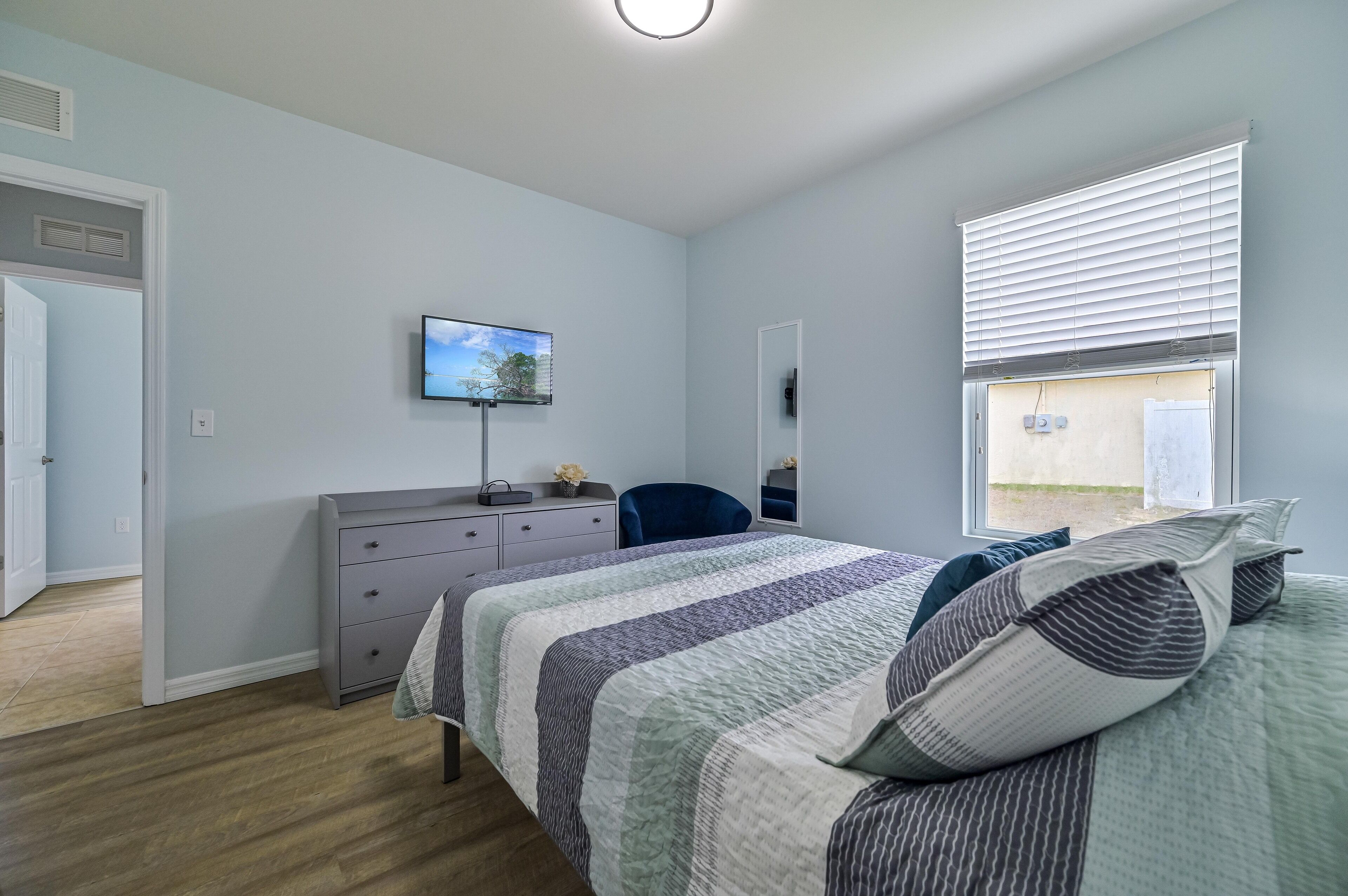 Un dormitorio con cama, cómoda, silla, televisor en la pared y ventana con persianas.