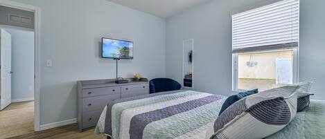 Un dormitorio con cama, cómoda, silla, televisor en la pared y ventana con persianas.