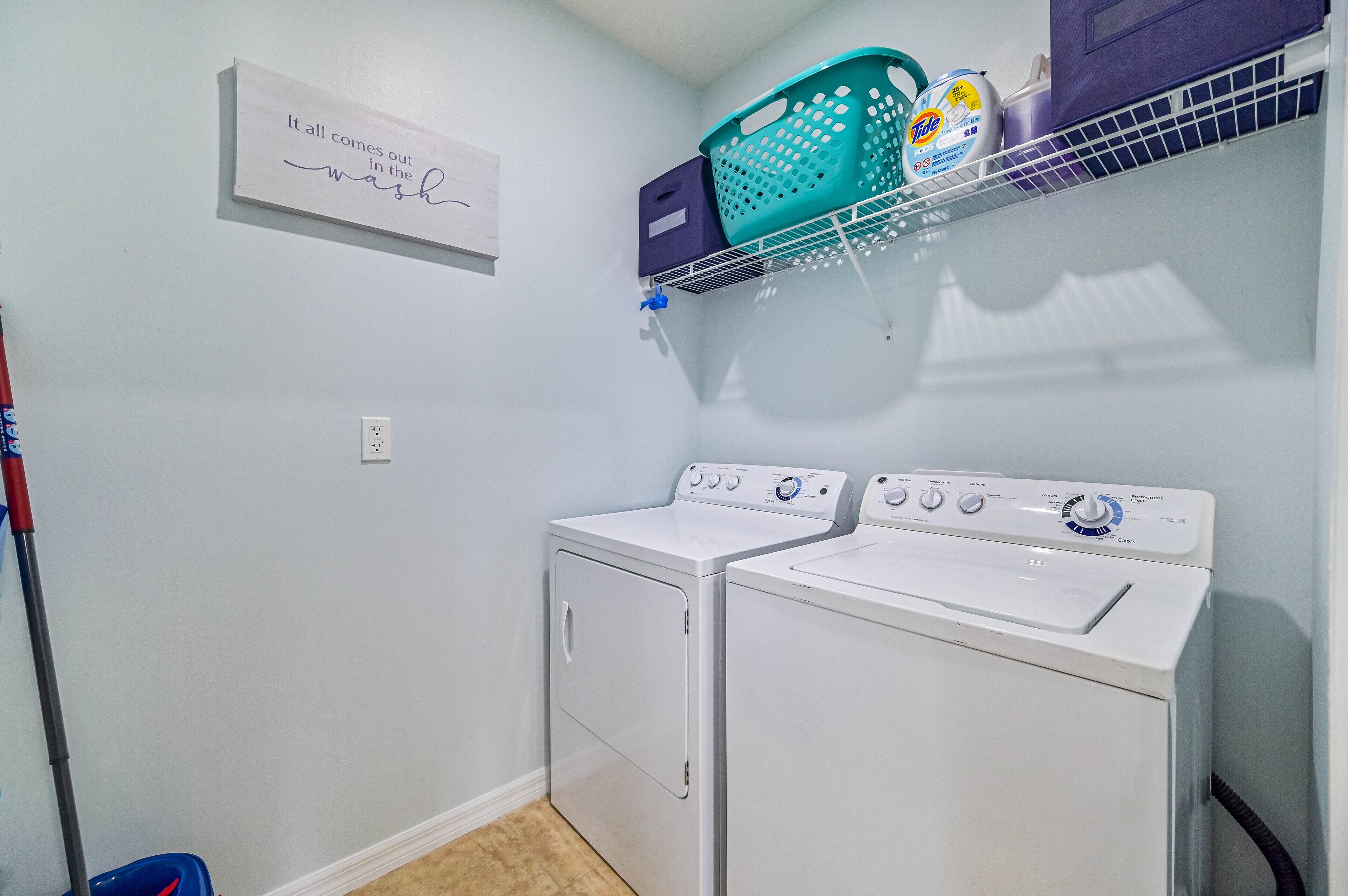 A laundry room with two white front-loading washing machines, a shelf with laundry detergent, and a wall-mounted sign.