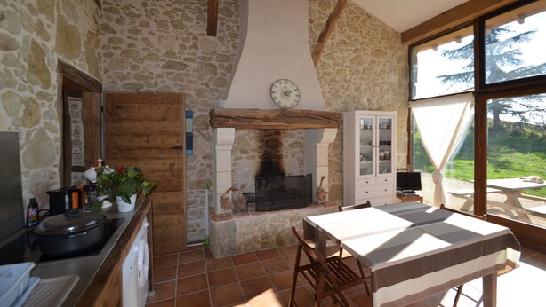 Une cuisine rustique avec un mur en pierre, des poutres en bois, une cheminée et un coin repas comprenant une table et des chaises.