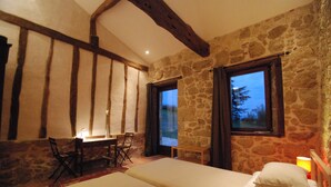 Une chambre avec un mur en pierre, un plafond en bois, un lit avec des oreillers, un bureau avec une chaise et une fenêtre donnant sur des arbres.