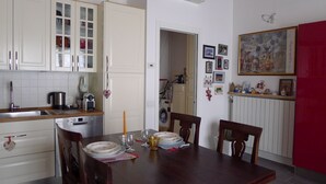 Ein Essbereich mit einem Holztisch, gedeckt für vier Personen, umgeben von Stühlen. Im Hintergrund ist eine Küche mit weißen Schränken und einer roten Tür erkennbar.