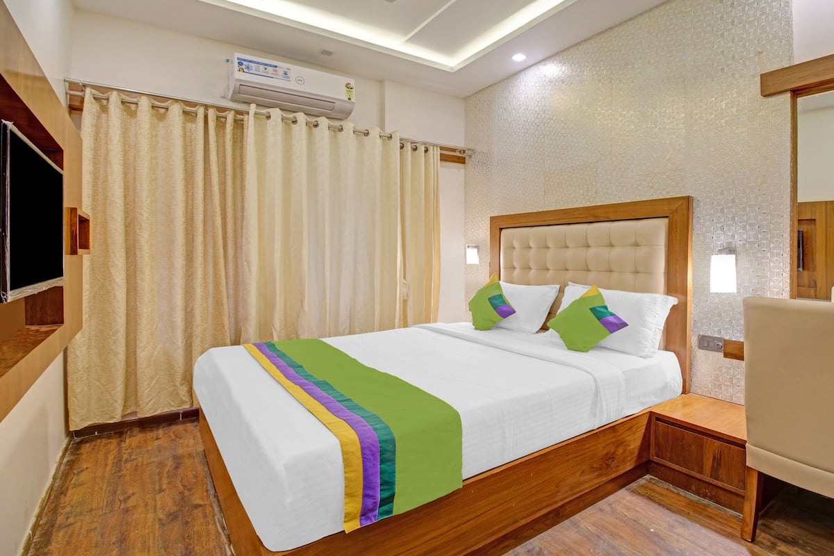 A hotel room with a bed, a wooden nightstand, a chair, a TV, and an air conditioning unit.