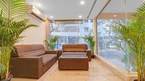 A modern living room with a brown sofa and ottoman, large windows with a view of the street, and several potted plants.
