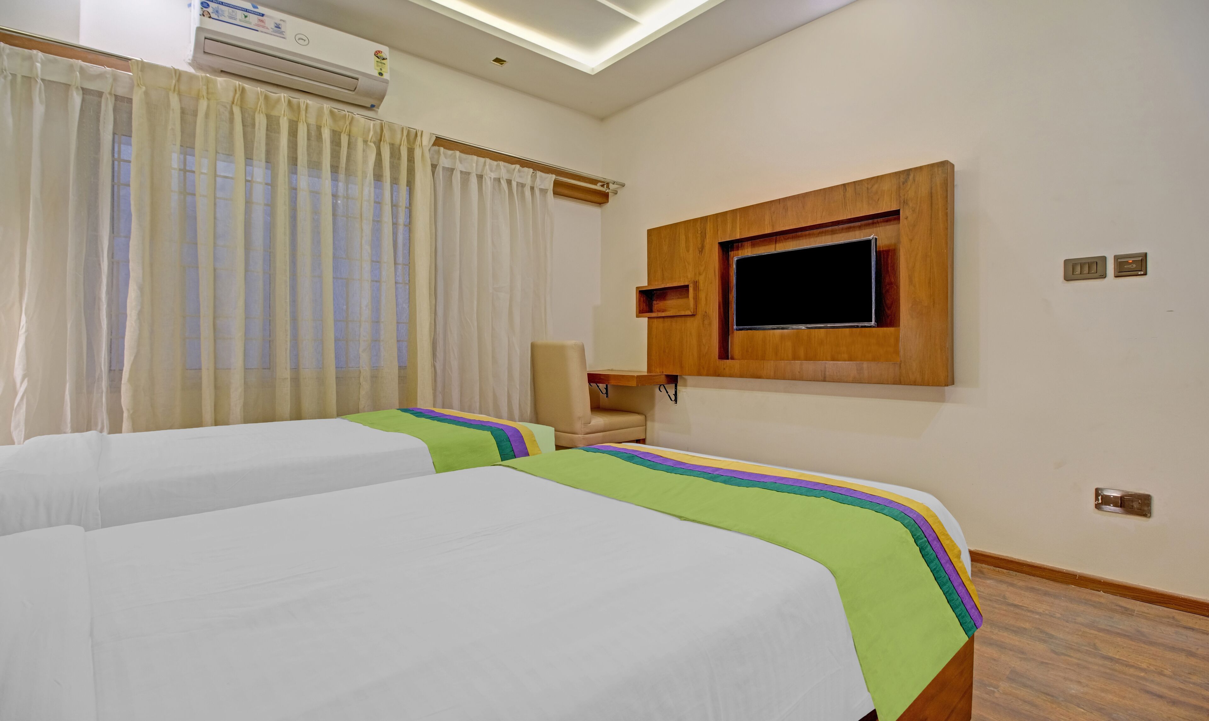 Una habitación de hotel con dos camas, un televisor montado en la pared, una silla y un equipo de aire acondicionado.