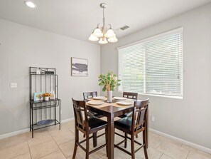 A dining area with a wooden table, chairs, a vase with flowers, a wall-mounted shelf with a sign, and a window with blinds.