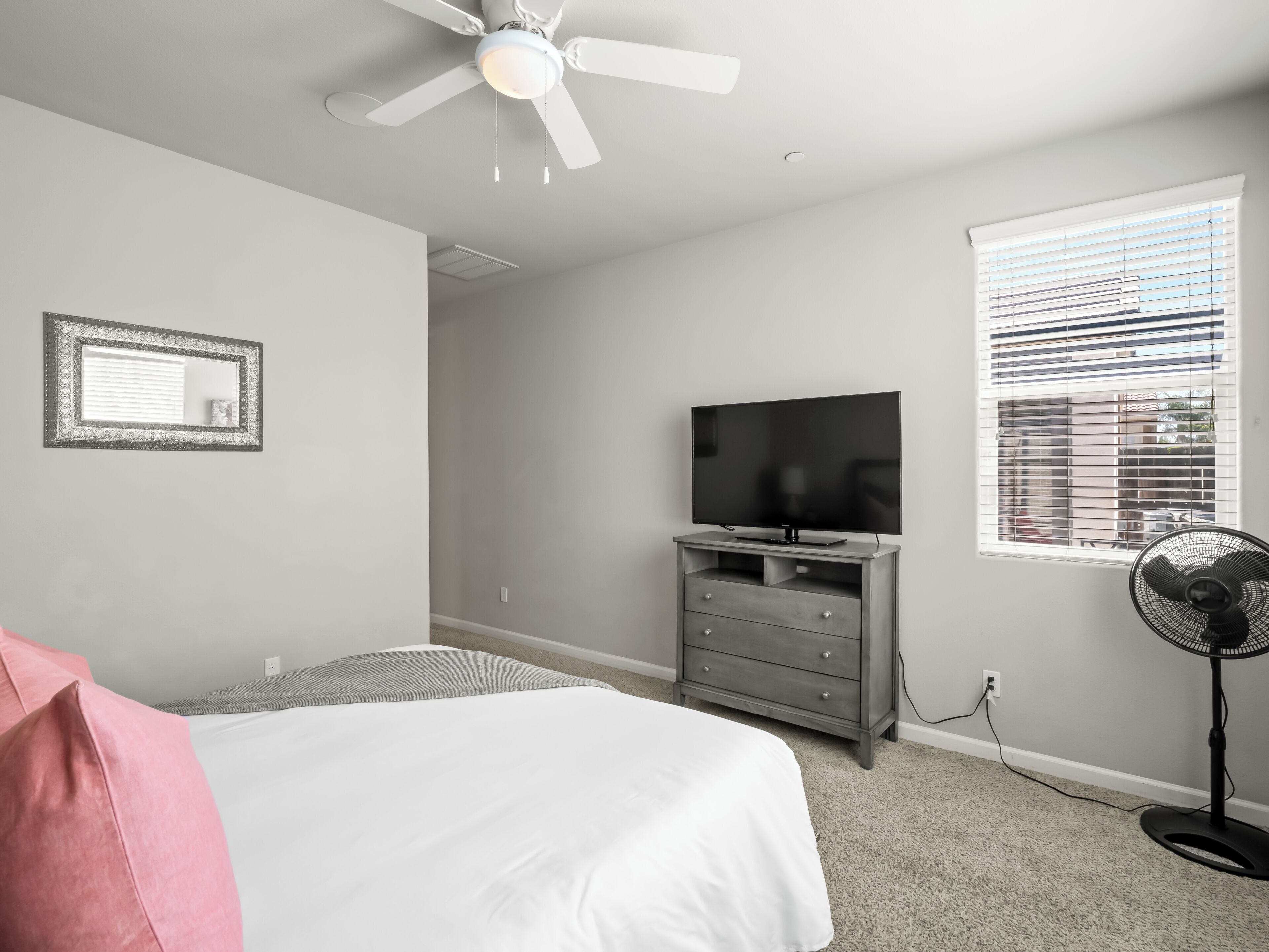 A bedroom with a bed, a television, a ceiling fan, a window with blinds, and a wall-mounted picture frame.