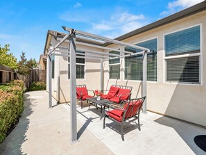 A covered patio area with a table and chairs, a pergola structure, and a view of a house with windows.