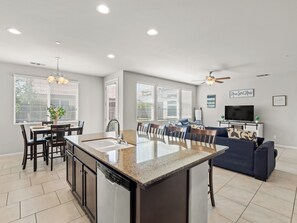 A modern kitchen with a central island, dining area with chairs, and a living room with a sofa and television. - Clovis House, Sleeps 10, Hammock, Covered Patio, Game Table (Clovis)