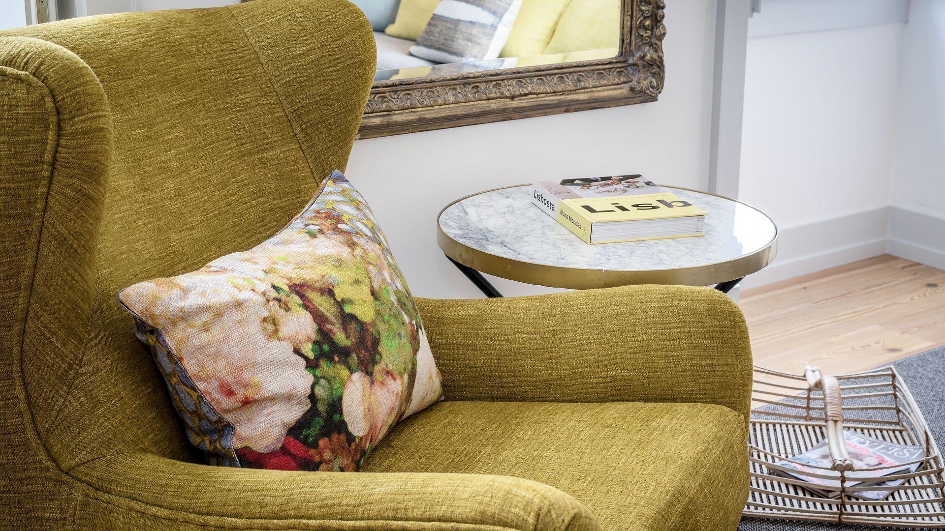 A mustard-colored armchair with a floral pillow, a small round table with a book titled 'Lisboeta', and a framed mirror on the wall.