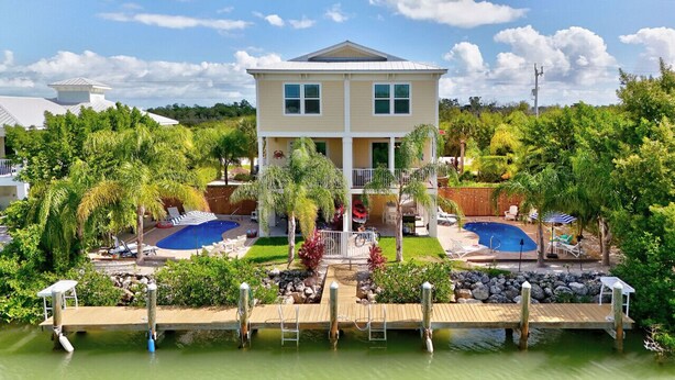 Una casa de dos plantas con un amplio balcón delantero, dos piscinas y jardines exuberantes.