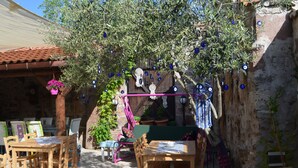 An outdoor seating area with wooden tables and chairs, a hanging flower pot, and a tree with blue decorative elements. - Nezisin Konagi (Ayvalik)