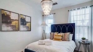 A bedroom with a bed, a chandelier, a lamp, a small table with a plant, and two abstract paintings on the wall. - Comfortable Chic | Long Term | Legacy (Plano)