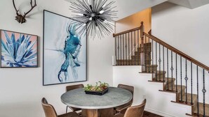 A dining area with a round table, four chairs, a chandelier, and a staircase with wooden railings. - Comfortable Chic | Long Term | Legacy (Plano)