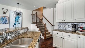 A kitchen with granite countertops, stainless steel sinks, white cabinetry, a staircase with wooden railings, and a coffee maker on the counter. - Comfortable Chic | Long Term | Legacy (Plano)