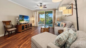 A spacious living room with hardwood floors, a ceiling fan, a large window with a view of palm trees, a sofa, and a coffee table.