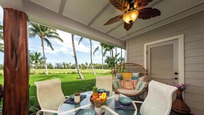 A covered outdoor seating area with a glass table, white chairs, and a wicker chair with cushions.