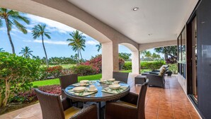 An outdoor patio with a round table set for dining, surrounded by wicker chairs, and overlooking a lush garden with palm trees and flowering plants. - Includes Beach Gear! Spacious lanai with resort views.  #2 Shores at Waikoloa (Waikoloa Beach Resort)