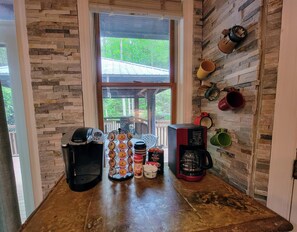 A kitchen area with a stone wall, a window with a view of greenery, and various coffee makers and mugs on the counter.