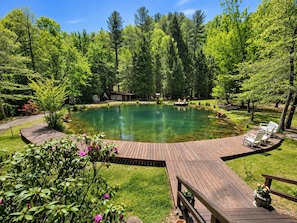 A wooden deck surrounding a clear pond with a small dock, surrounded by lush greenery and trees. - Pet-Friendly Waterfront² Lodge | Hot Tub, Fire Pit & Game Room (Cherry Log)