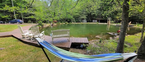 A hammock on a wooden deck overlooking a pond, surrounded by trees and a small shed.