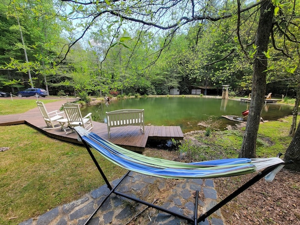 A hammock on a wooden deck overlooking a pond, surrounded by trees and a small shed.