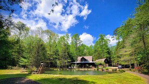 Eine rustikale Holzhütte mit Teich, umgeben von dichtem Wald und blauem Himmel.