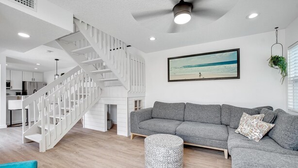A spacious living area with a grey sofa, a round ottoman, a white staircase, and a ceiling fan. - Beachside Duplex A (Saint Augustine Beach)