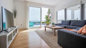 Ein modernes Wohnzimmer mit großem Fernseher, einer Couch, einem Couchtisch und Blick auf das Meer durch große Schiebetüren.