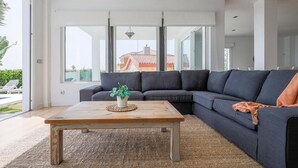 Ein modernes Wohnzimmer mit einem dunkelgrauen Ecksofa, einem Holztisch mit Pflanze und großen Fenstern mit Blick auf ein Haus und Grünflächen.